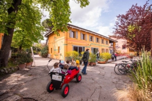 Children in a red cart outside the Kilowatt building at Le Serre dei Giardini Margherita, a green urban hub in Bologna.